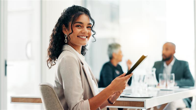 A woman looking up and smiling from her tablet as she works in a modern, sunlight-filled office.