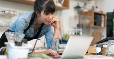Business owner of a cafe leaning over the counter and looking at a laptop.