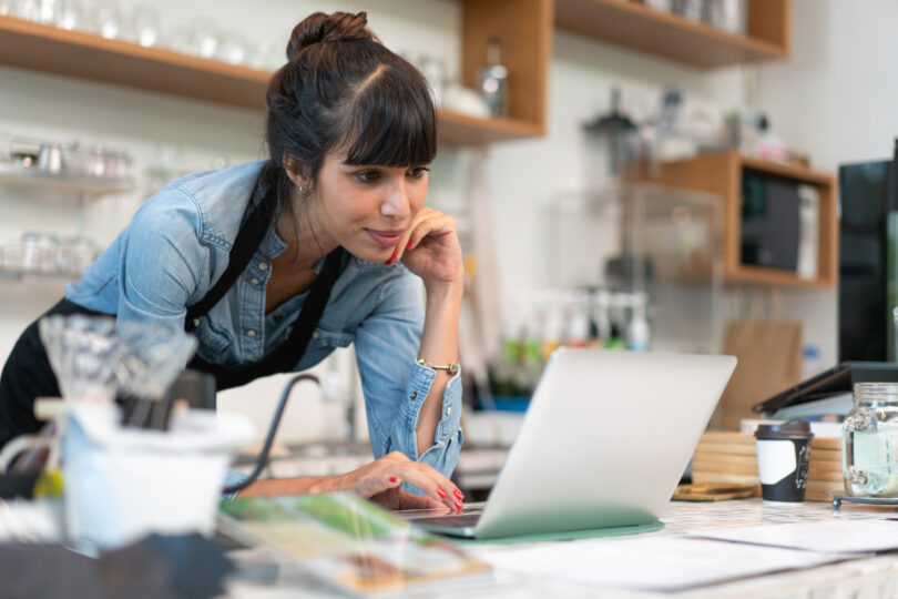 Business owner of a cafe leaning over the counter and looking at a laptop.