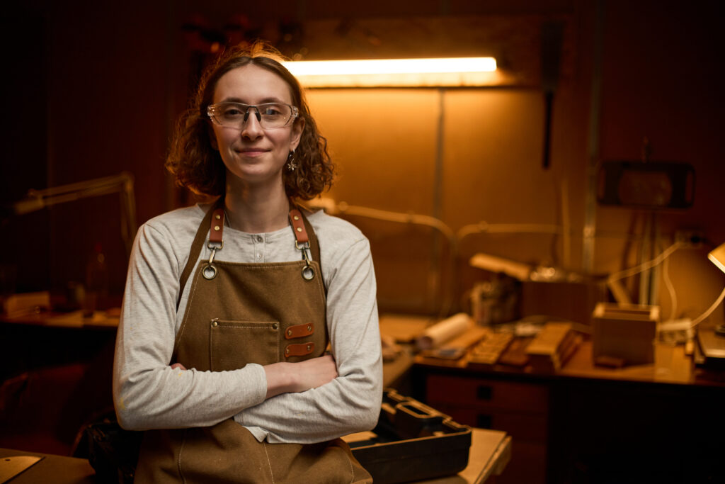 Portrait of young adult Caucasian woman carpenter standing with crossed arms in carpentry workshop, representing skilled trades and small business branding. Medium shot